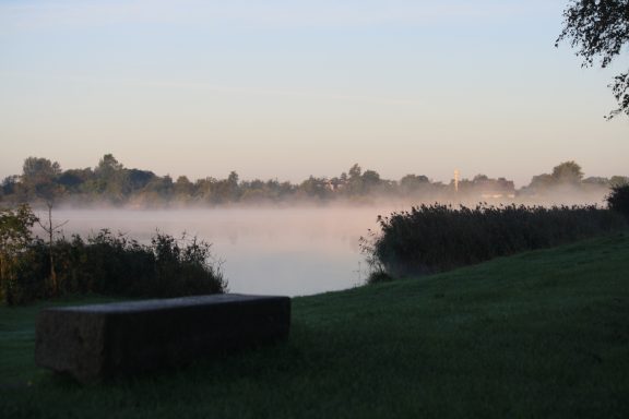 Ruhiger Fluß mit Nebel und Bäumen im Hintergrund bei Sonnenaufgang.