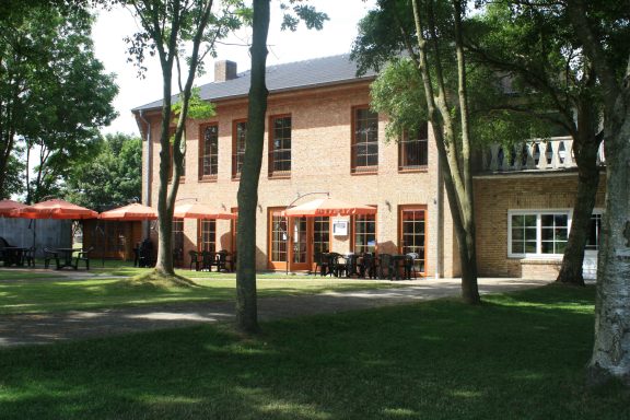 Gelbes Backsteingebäude mit Terrassen unter Bäumen, umgeben von Grünflächen.