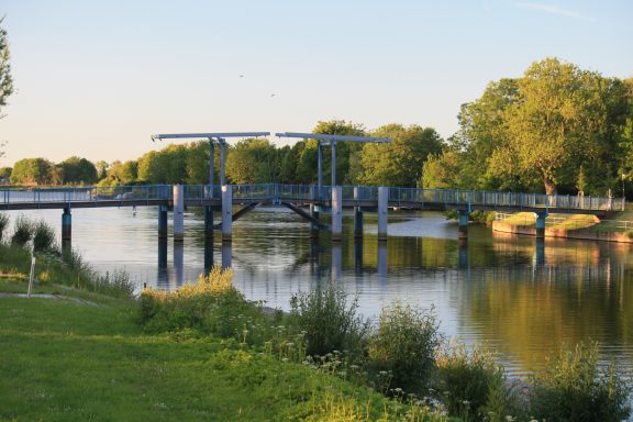 Blaue Fußgängerbrücke in Friedrichstadt über die ruhige Treene, umgeben von grüner Vegetation.