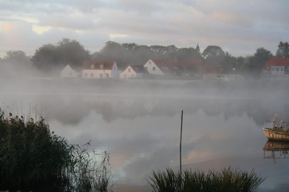 Nebel über der ruhigen Treene mit weißen Häusern aus Friedrichstadt im Hintergrund.